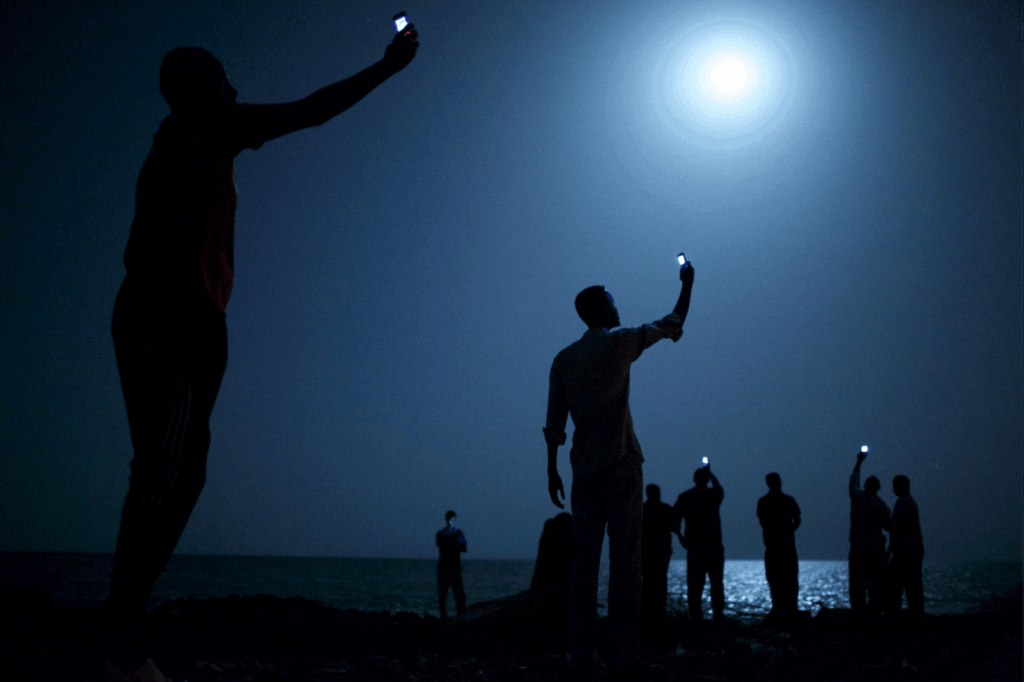 Searching for a Signal: Migrants in Djibouti Catching Cell Signal at Night. «Signal». Photo: John Stanmeyer. Image source: nationalgeographic.com. Searching for a Signal: Migrants in Djibouti Catching Cell Signal at Night. «Signal». Photo: John Stanmeyer. Image source: nationalgeographic.com. Photo.