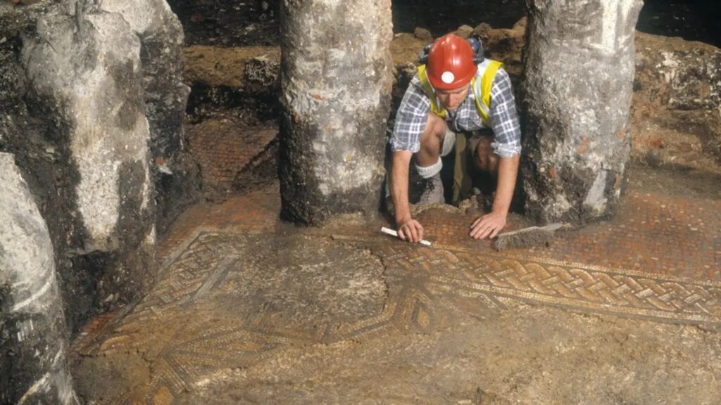 An archaeologist excavates a mosaic floor with a complex geometric pattern in a late Roman extension. Photo.
