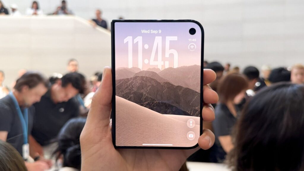 A folded iPhone Duo in hand: the outer screen shows the clock, with the front camera cutout in the top-right corner