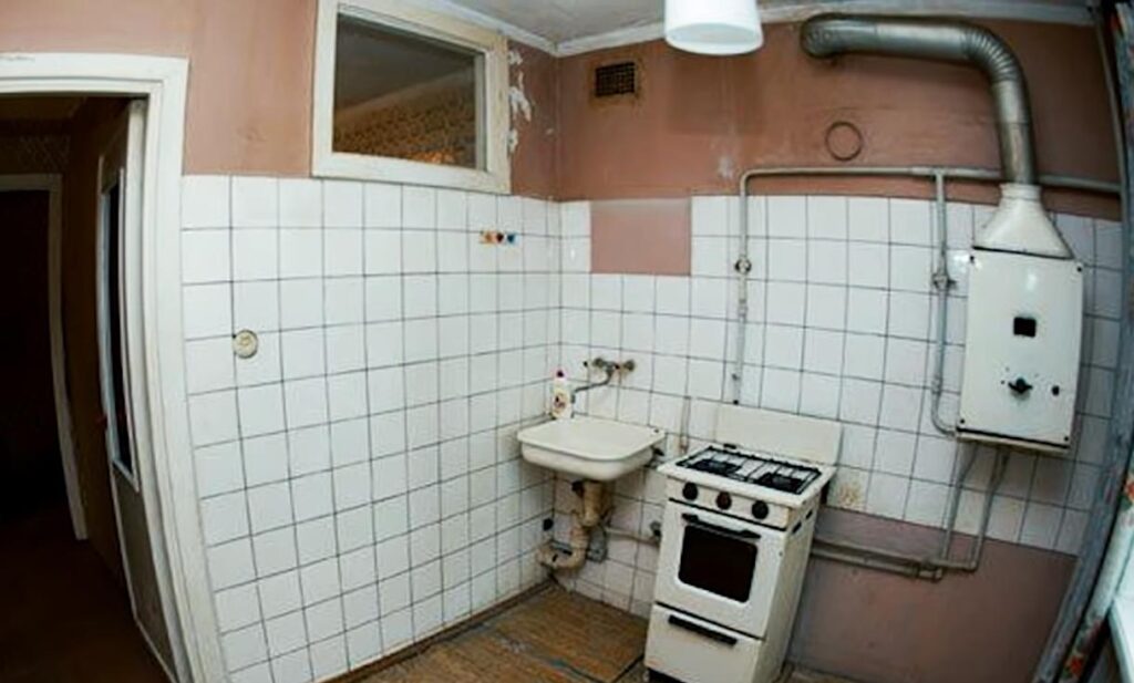 An old Soviet kitchen with a gas water heater, exposed pipes and a small window into the adjoining room.